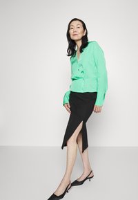 Blusa verde com bolinhas, com fecho frontal de botões, combinada com uma saia preta com uma fenda alta na coxa, usada com sapatos de salto tipo slingback pretos.