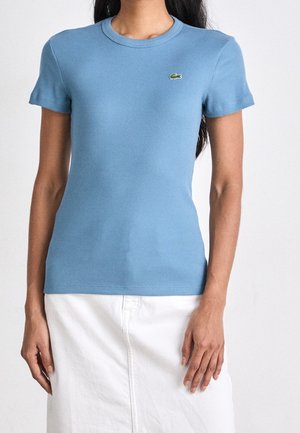 Lichtblauw geribd T-shirt met korte mouwen, ronde halslijn en een klein groen logo. De stof is rekbaar en sluit nauw aan. Draagt bij een witte rok.