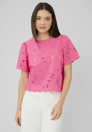 Femme aux cheveux bruns raides portant un haut en broderie anglaise rose vif à motif floral et un pantalon blanc, sur un fond uni gris clair.