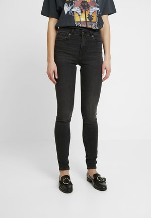 Jean skinny noir avec une texture lisse, présentant une taille mi-haute et un design classique à cinq poches. Associés à des mocassins noirs avec un accent doré.