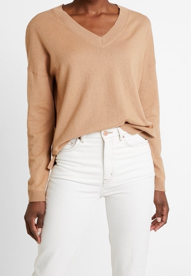 Femme portant un pull camel à col en V et manches longues, rentré dans un jean blanc taille haute, debout avec les bras détendus le long du corps.