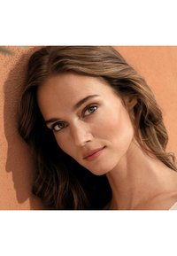 Portrait d'une personne avec de longs cheveux bruns ondulés, une peau lisse et des yeux verts sur un fond orange texturé. Un éclairage naturel met en valeur les traits.