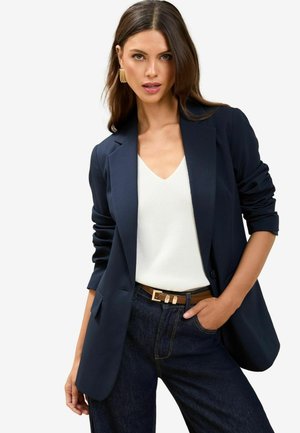 Navy blazer with tailored design, lightweight fabric, and single button closure, paired with a white v-neck top and dark denim jeans.
