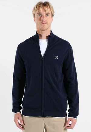 Pull zippé bleu marine en tissu tricoté doux, avec un col montant, des poignets côtelés et un petit logo sur le côté supérieur gauche.