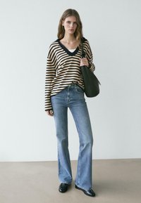 Žena nosící béžový a černý pruhovaný svetřík s výstřihem do V, světle modré džíny s vysokým pasem, černé boty, drží na rameni velkou černou kabelku.