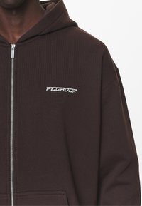 Felpa con zip marrone, dotata di tasca frontale, realizzata in tessuto morbido, con un logo argento "PEGADOR" sul petto.