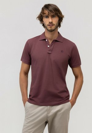 Hombre con cabello castaño y barba, vistiendo una camiseta polo granate y pantalones beige, de pie con las manos en los bolsillos frente a un fondo liso.