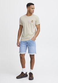 Beige bomulds t-shirt med et farverigt cirkulært logo, parret med lyseblå denim shorts med opfoldede kanter, og brune sneakers med gule accenter.