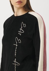 Donna che indossa un top nero a maniche lunghe con strisce bianche e mauve sulle maniche e testo corsivo verticale sul davanti.