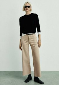 Maglione nero con polsini bianchi, jeans beige a gamba larga, cintura nera e stivaletti. La modella indossa occhiali da sole neri su uno sfondo neutro.
