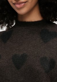 Καφέ μαλακό πουλόβερ με φουντωτή υφή, διακοσμημένο με μαύρα σχέδια καρδιών. Υψηλή λαιμόκοψη και διακριτικές, άνετες λεπτομέρειες σχεδίασης.