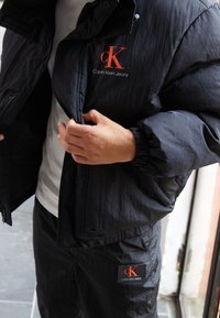 Svart puffjacka med dragkedja och ribbade ärmslut, med röd "Calvin Klein Jeans" logotyp på bröstet och matchande träningsoverallbyxor med logotygmärke.