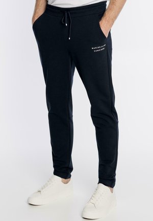 Mężczyzna w czarnych spodniach dresowych z białym logo "Marine Club Campione" i białych trampkach, z rękami w kieszeniach, stojący na białym tle.