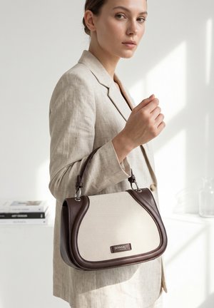 Donna con blazer beige che tiene una borsa beige e marrone con manico superiore, in piedi in una stanza minimalista ben illuminata.