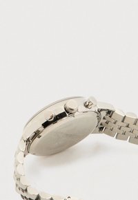 Relógio de pulso em aço inoxidável prateado mostrando o fundo da caixa, botões laterais e pulseira de metal ligada sobre um fundo claro.