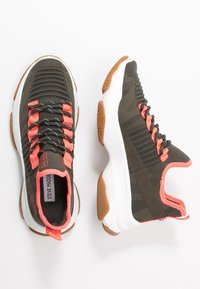 Tummanvihreät urheilukengät, joissa on teksturoitu neulottu päällinen, oranssit yksityiskohdat, mustat nauhat ja valkoinen välipohja. Kumipohja tarjoaa pitoa.