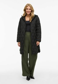 Black quilted long coat with a hood, featuring a zip front, diamond pattern, and side pockets, paired with dark green wide-leg pants.