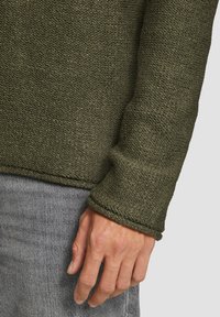 Olive green textured sweater o luźnym kroju, z zaokrąglonym dołem i długimi rękawami. Szare dżinsy częściowo widoczne, z ręką opartą na nich.