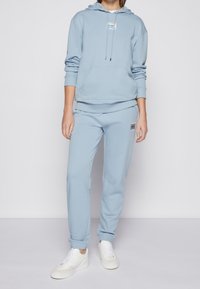 Sudadera azul clara con un bolsillo frontal, combinada con pantalones de chándal a juego. Presenta un parche con el logo. Zapatillas blancas completan el atuendo.