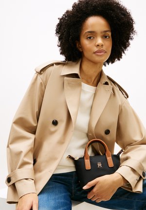 Young woman with curly hair wearing a beige trench coat, white sweater, blue jeans, holding a small black handbag with brown handles.