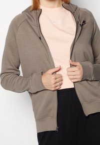 Ljusbrun zip-up hoodie med en mjuk textur, som har en huva, framfickor och en svart Nike-logga på bröstet, bärs över en ljusrosa t-shirt.