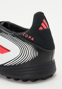 Schwarzer Fußballschuh mit weißen und roten Akzenten, ausgestattet mit einem strukturierten Obermaterial, markanten geschwungenen Streifen und einer gepolsterten Ferse mit Branding.