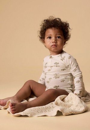 Tout-petit aux cheveux bouclés assis sur une couverture beige en tricot, portant un body à manches longues de couleur crème avec des imprimés d'animaux et d'arbres.