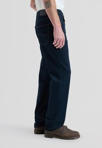 Vyras dėvi tamsiai mėlynas džinsus, baltą marškinius, rudas odines batus, stovi šonu, rodo tatuiruotes ant dilbio ir rankos, kuri yra atsipalaidavusi šalia.