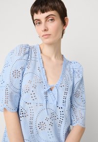 Lichtblauw geborduurd blouse met eyelet uitsparingen, korte pofmouwen, een gerimpelde halslijn en een strikdetail op de borst.