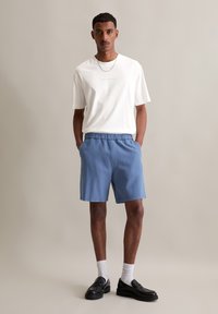 Witte T-shirt met gecentreerde tekst, lichtblauwe shorts met elastische tailleband, en zwarte instappers, aangevuld met witte crew-sokken.