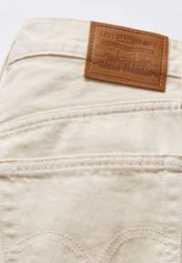 Tissu denim beige avec une poche arrière cousue et une étiquette en cuir marron Levi Strauss & Co. cousue à la ceinture.