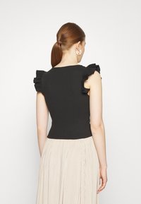 Top preto canelado com mangas com babados, design ajustado e decote redondo. Usado com uma saia plissada beige, mostrando um contraste têxtil suave.