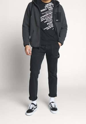 Szara bluza polarowa na zamek, nałożona na czarną koszulkę z nadrukiem, w zestawie z czarnymi dżinsami i czarno-białymi trampkami. Minimalne detale na spodniach.
