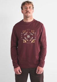 T-shirt manches longues bordeaux en coton, avec un motif graphique en or et argent portant les inscriptions « STAY ALIVE » et « SINCE 1995 ».