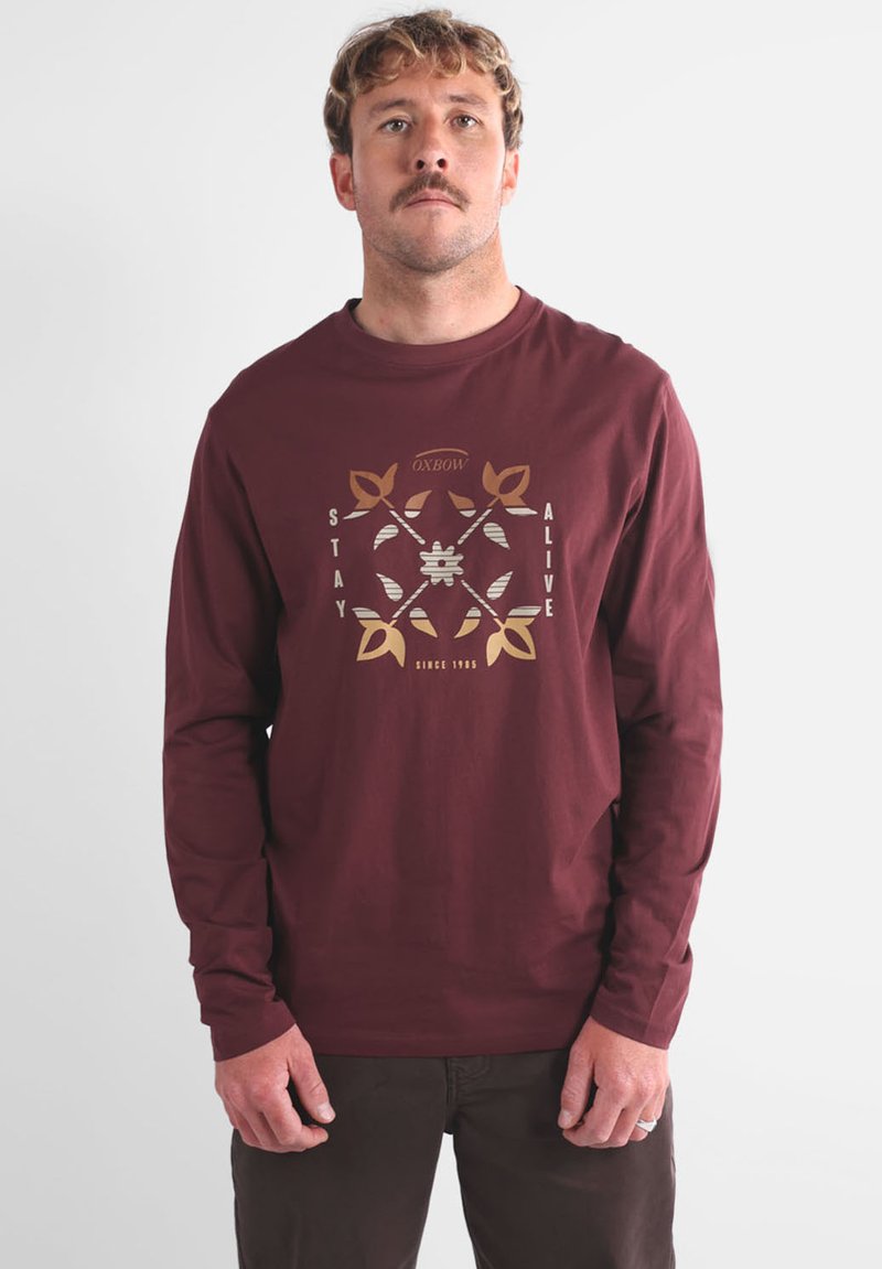 T-shirt manches longues bordeaux en coton, avec un motif graphique en or et argent portant les inscriptions « STAY ALIVE » et « SINCE 1995 ».