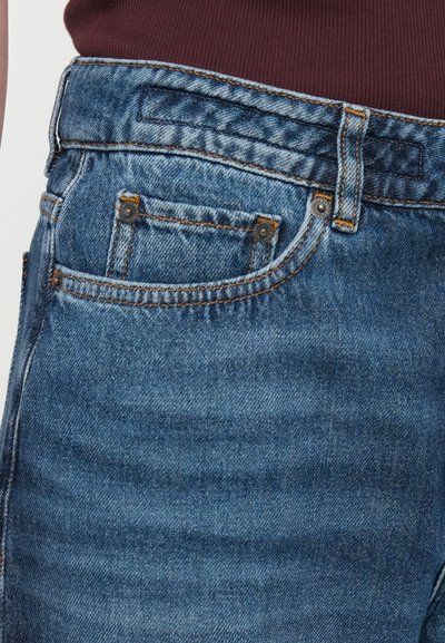 Jean en denim de couleur bleu moyen avec un léger délavage. Présente des poches avant, des coutures orange contrastantes et des accents métalliques.