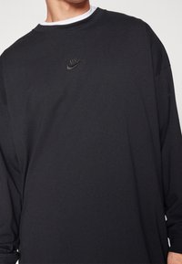 Svart långärmad t-shirt med rund halsringning, med en subtil svart Nike-logotyp på bröstet. Tyget verkar mjukt med en slät yta.