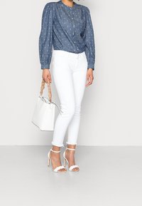 Džínová košile na knoflíky s nabíranými rukávy, spárovaná s bílými skinny džíny. Bílá kabelka s pleteným uchopem a bílé sandály na vysokém podpatku.