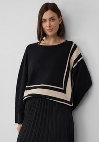 Schwarzer Pullover mit creme- und schwarzen geometrischen Mustern. Er hat eine lockere Passform, einen runden Halsausschnitt und gerippte Bündchen. Kombiniert mit einem plissierten schwarzen Rock.