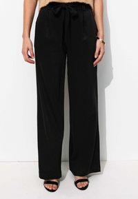 Pantalon large noir en tissu lisse, avec une taille élastique ornée d'un nœud. Accompagné de sandales noires à lanières.