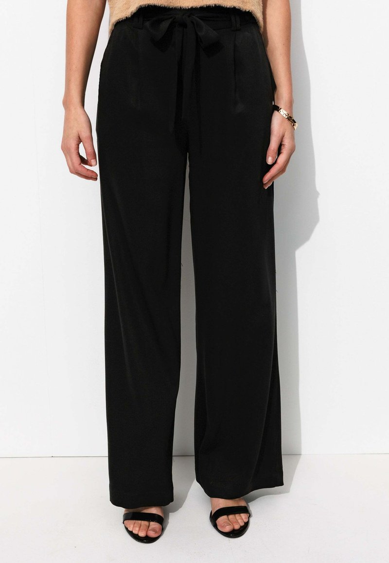 Pantalon large noir en tissu lisse, avec une taille élastique ornée d'un nœud. Accompagné de sandales noires à lanières.