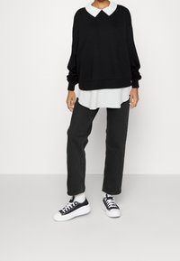 Maglione nero oversize con colletto bianco indossato sopra una tunica bianca. Jeans neri a gamba dritta e sneakers nere con suola bianca.