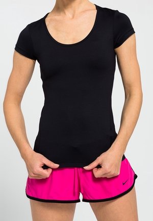 Person trägt ein eng anliegendes schwarzes Kurzarmshirt und leuchtend pinke Sportshorts und steht mit den Händen am Saum des Shirts.