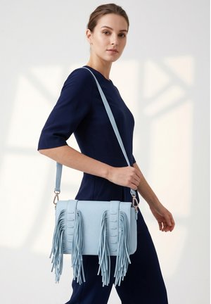 Femme en robe bleu marine portant un sac à bandoulière bleu clair avec de longues franges, marchant devant un fond neutre.