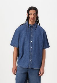 Cămașă din denim cu mâneci scurte, culoare albastru deschis, închidere cu nasturi și croială lejeră. Textură moale cu detalii de cusătură minime.