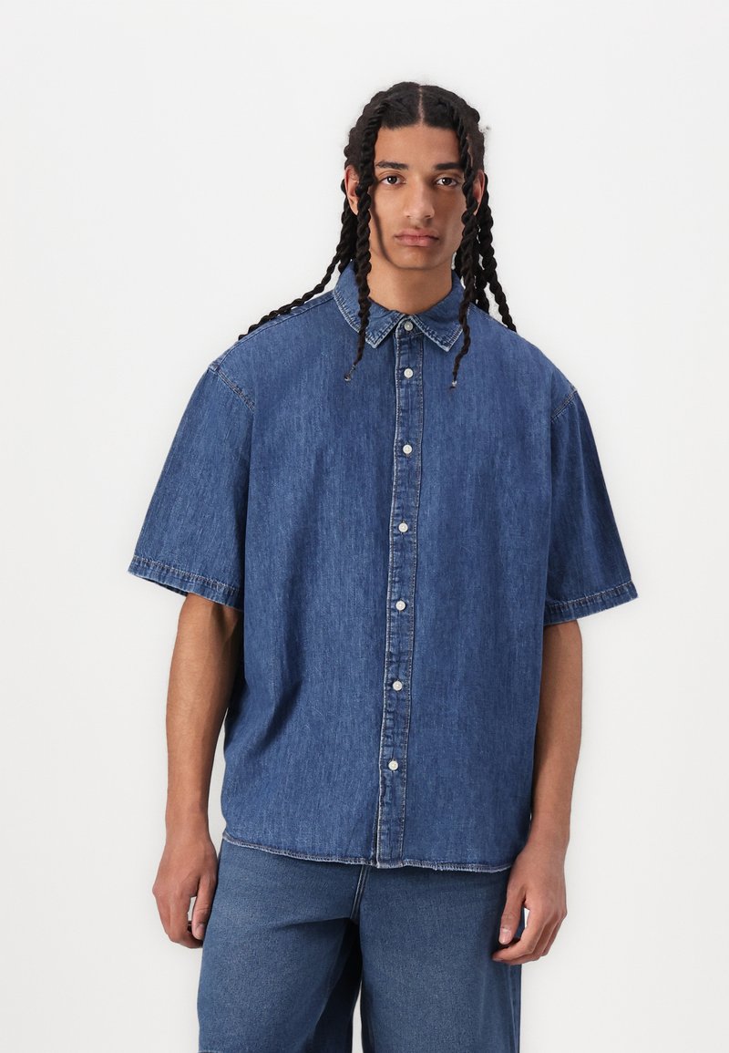 Cămașă din denim cu mâneci scurte, culoare albastru deschis, închidere cu nasturi și croială lejeră. Textură moale cu detalii de cusătură minime.