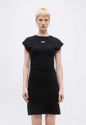 Lühikeste blondide juustega naine kannab kehast lähedalt järgivat musta kleiti, millel on rinnal "GAP" logo, seistes ühevärvilise hele tausta ees.