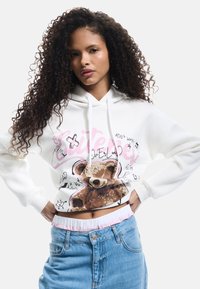 Witte hoodie met een teddybeerafbeelding en kleurrijke doodles. De mouwen zijn geribbeld en de tailleband is elastisch met een roze accent.