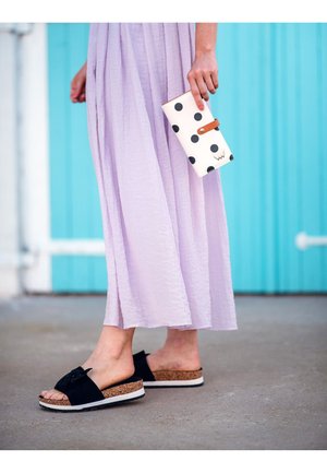 Black slide sandals with a bow, cork sole, and white accent stripes paired with a lavender, flowy skirt and a white wallet with black polka dots.