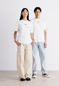 To modeller står sammen iført hvite bomull t-skjorter med en liten logo. Den ene har på seg beige løse bukser, mens den andre har lyseblå jeans.
